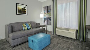 Ein Hotelzimmer mit grauem Sofa, blauem Hocker, einem kleinen Tisch, einer Wandleuchte, einem großen Fenster mit Vorhängen und einem gerahmten Bild an der Wand.