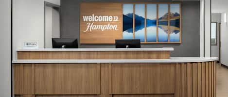 A hotel reception desk with a wooden counter, a "Welcome to Hampton" sign, and a scenic wall art piece.