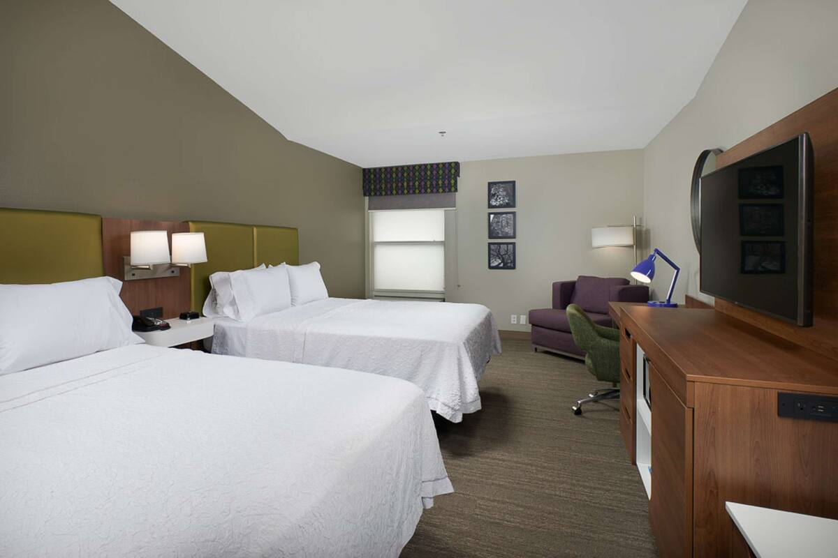 A hotel room with two beds, a desk with a television, a chair, and a window with blinds.