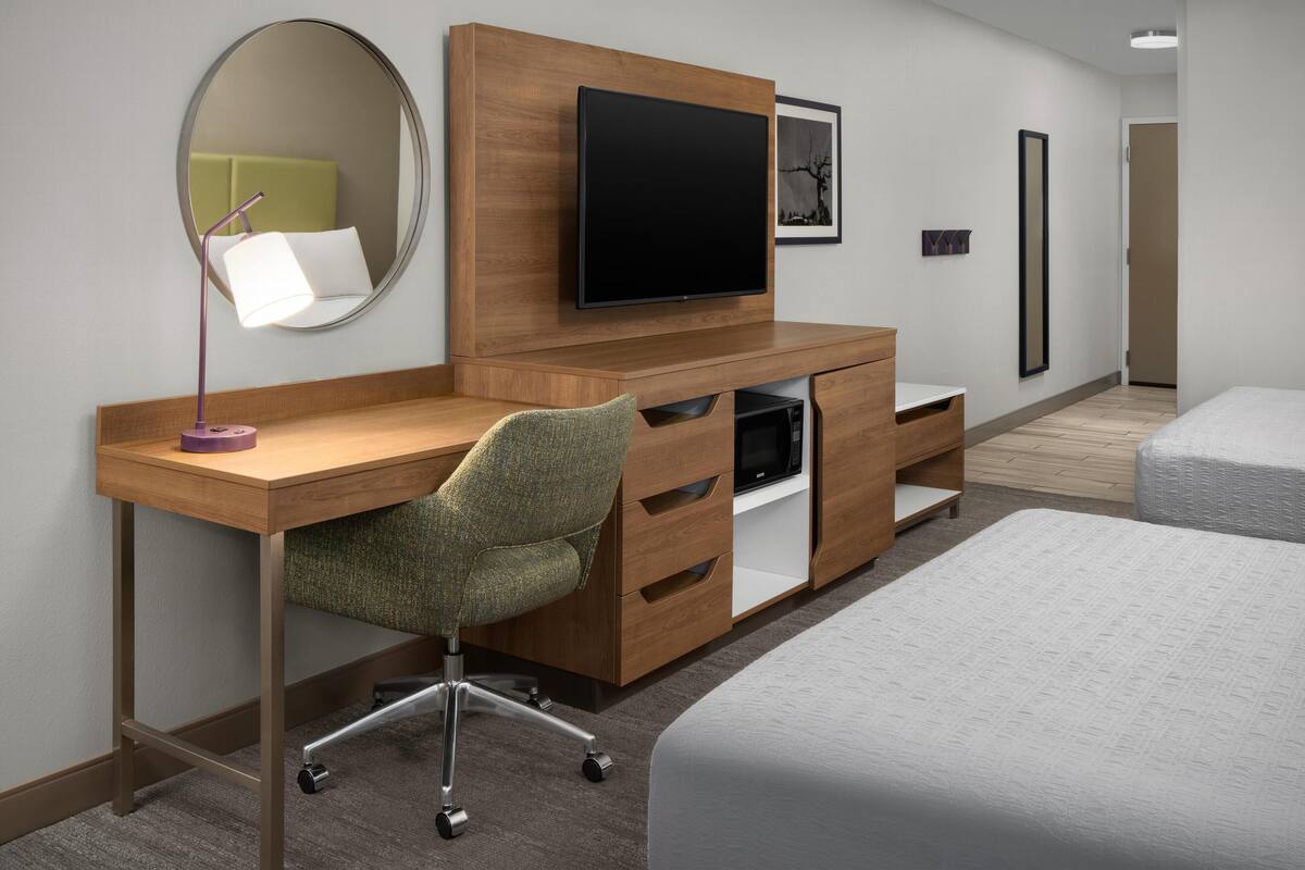 A modern hotel room with a wooden desk, a chair, a television, a bed, and a mirror.