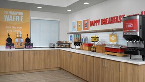 A modern kitchen area with a breakfast bar, waffle maker, juice dispenser, and various breakfast items.