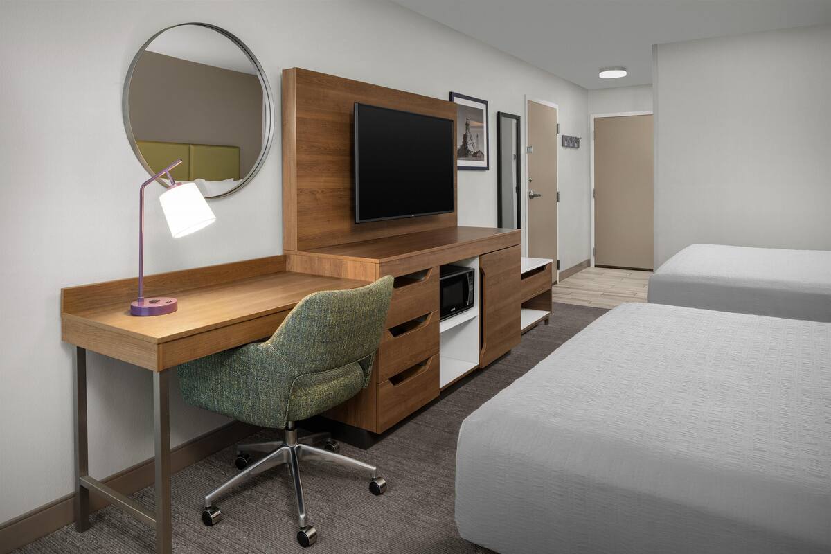 A hotel room with a wooden desk, a green office chair, a flat-screen TV, a bed, and a door.