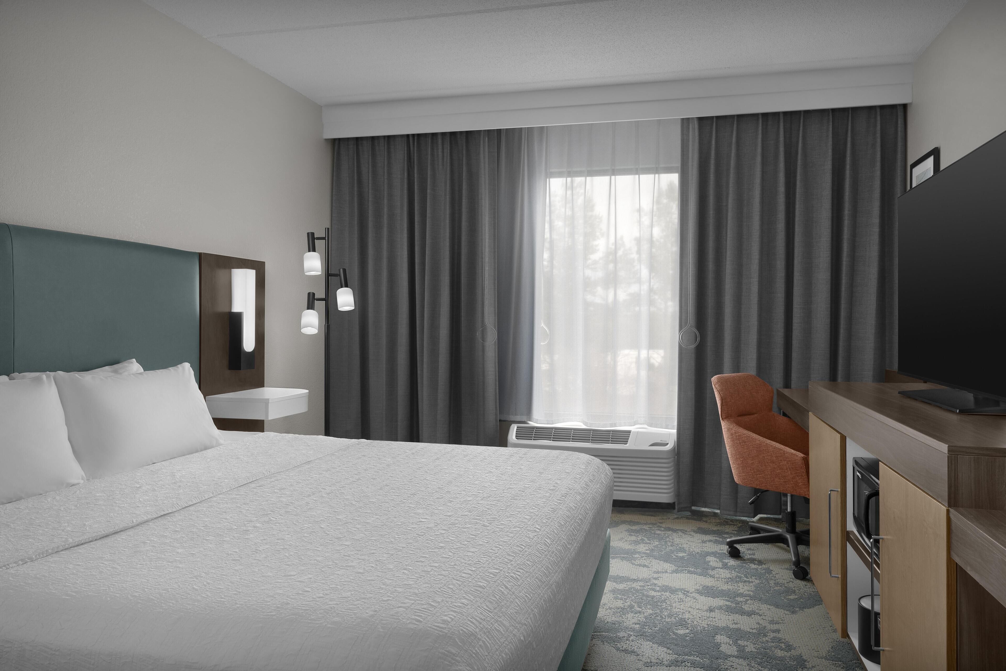 Une chambre d’hôtel avec un grand lit, un bureau avec une chaise, un téléviseur à écran plat, une fenêtre avec des rideaux et un luminaire fixé au mur.