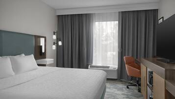 Une chambre d’hôtel avec un grand lit, un bureau avec une chaise, un téléviseur à écran plat, une fenêtre avec des rideaux et un luminaire fixé au mur.