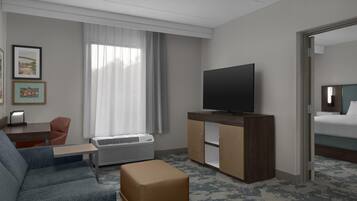 Habitación de hotel con sofá, televisión sobre un atril de madera, cama, ventana con cortinas y suelo alfombrado.