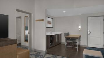 A hotel room with a television, a sofa, a dining area with a table and chairs, a kitchen area with a sink, and a door.