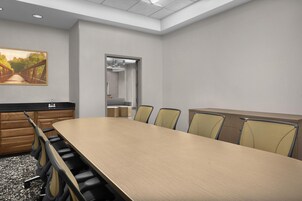 Un salón de conferencias con una larga mesa, sillas, un cuadro y vistas a través de una puerta.