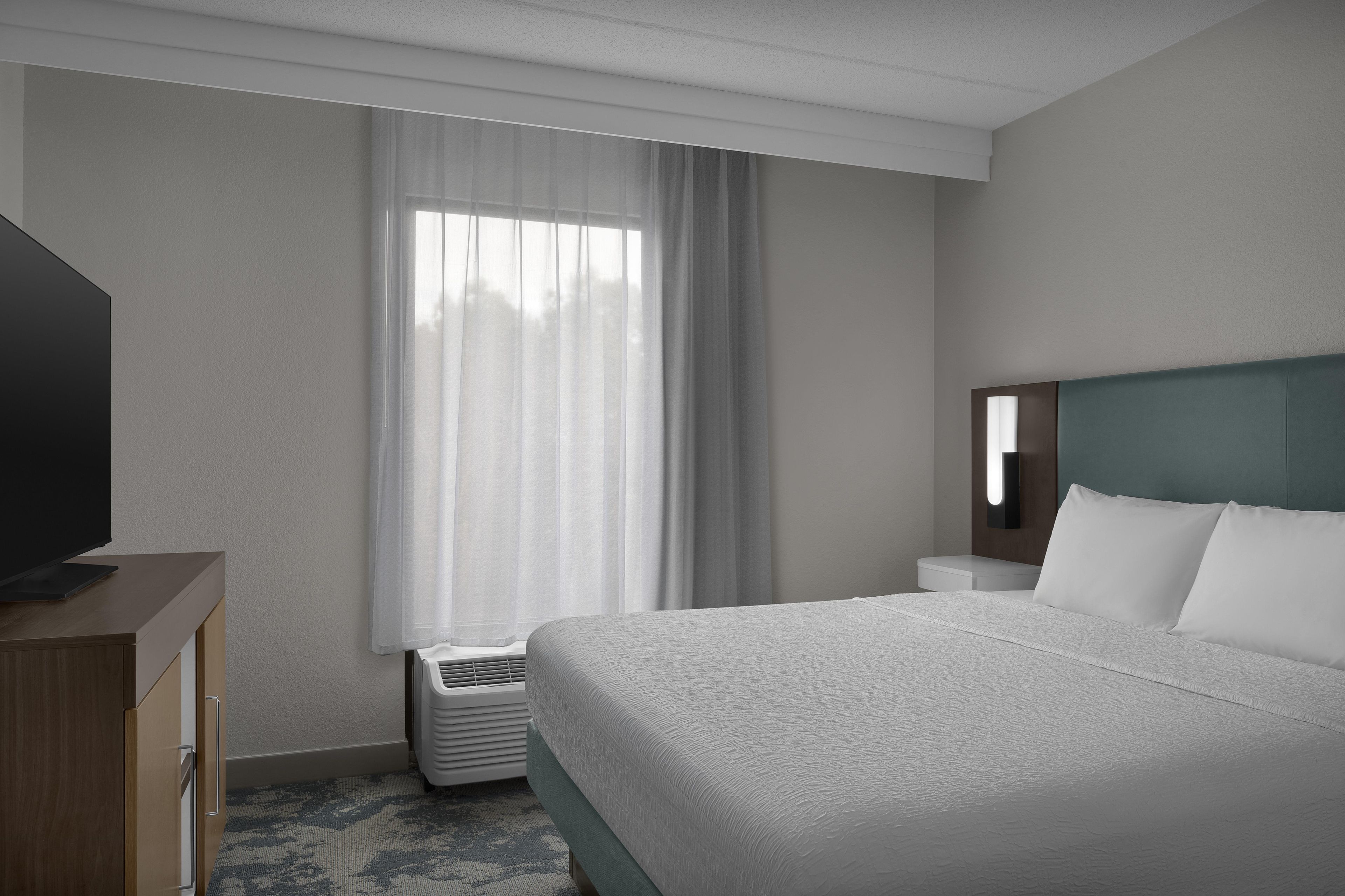 A hotel room with a bed, a television, a window with curtains, and a wall-mounted light fixture.