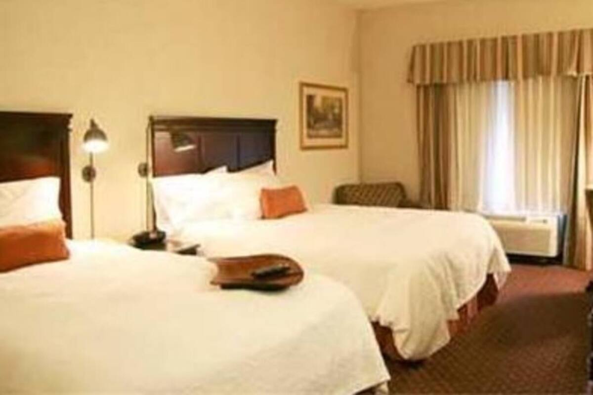 A hotel room with two beds, a nightstand, a lamp, a framed picture on the wall, and a window with curtains.