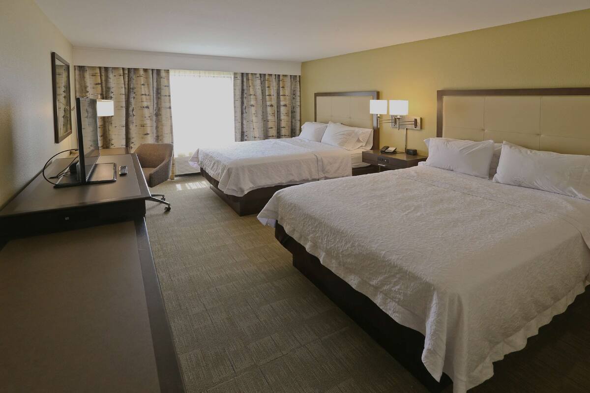 A hotel room with two beds, a desk with a computer, a chair, and a window with curtains.