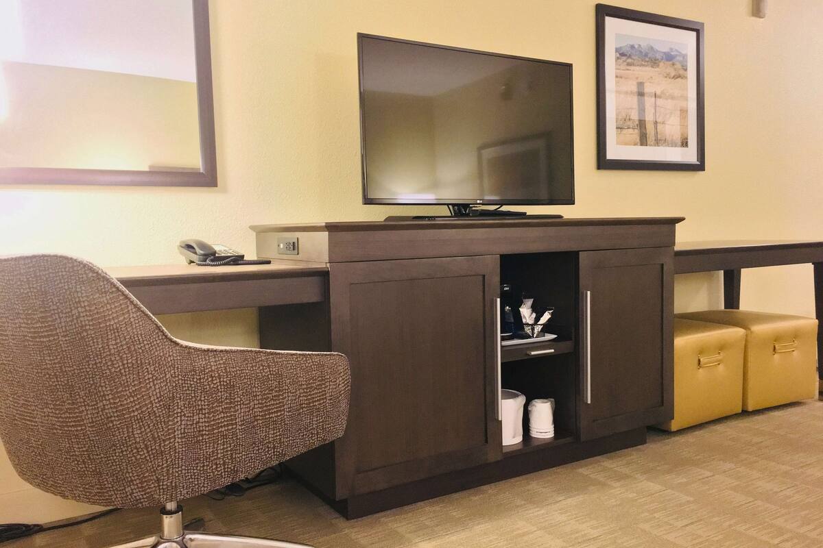 A hotel room with a desk, a chair, a television, a framed picture on the wall, and a small cabinet with drawers.