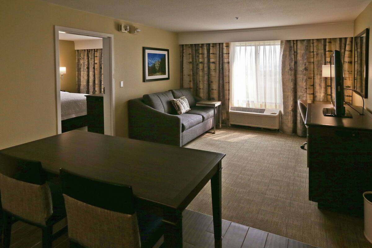 A hotel room with a dining table, a sofa, a TV, and a window with curtains.