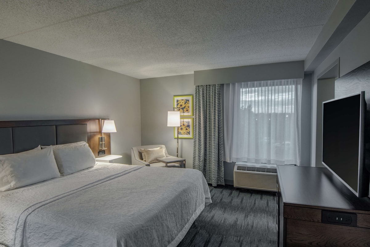 A hotel room with a large bed, a desk with a flat-screen TV, a chair, a lamp, and a window with curtains.