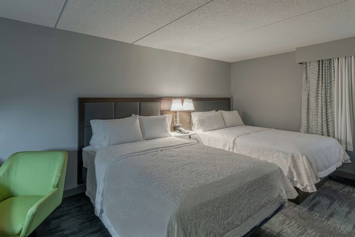 A hotel room with two beds, a green chair, a nightstand with a lamp, and a window with curtains.