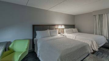 Une chambre d’hôtel avec deux lits, un fauteuil vert, une table de chevet avec une lampe et une fenêtre avec des rideaux.