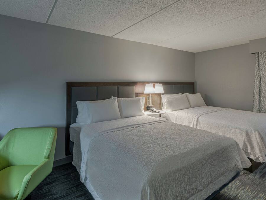 Une chambre d’hôtel avec deux lits, un fauteuil vert, une table de chevet avec une lampe et une fenêtre avec des rideaux.