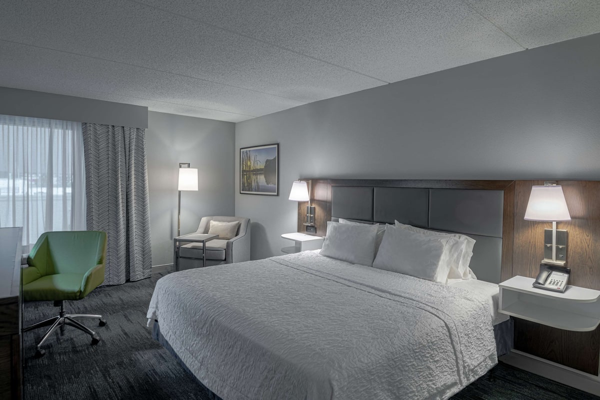 A hotel room with a large bed, two bedside tables with lamps, a green chair, a desk, and a window with curtains.