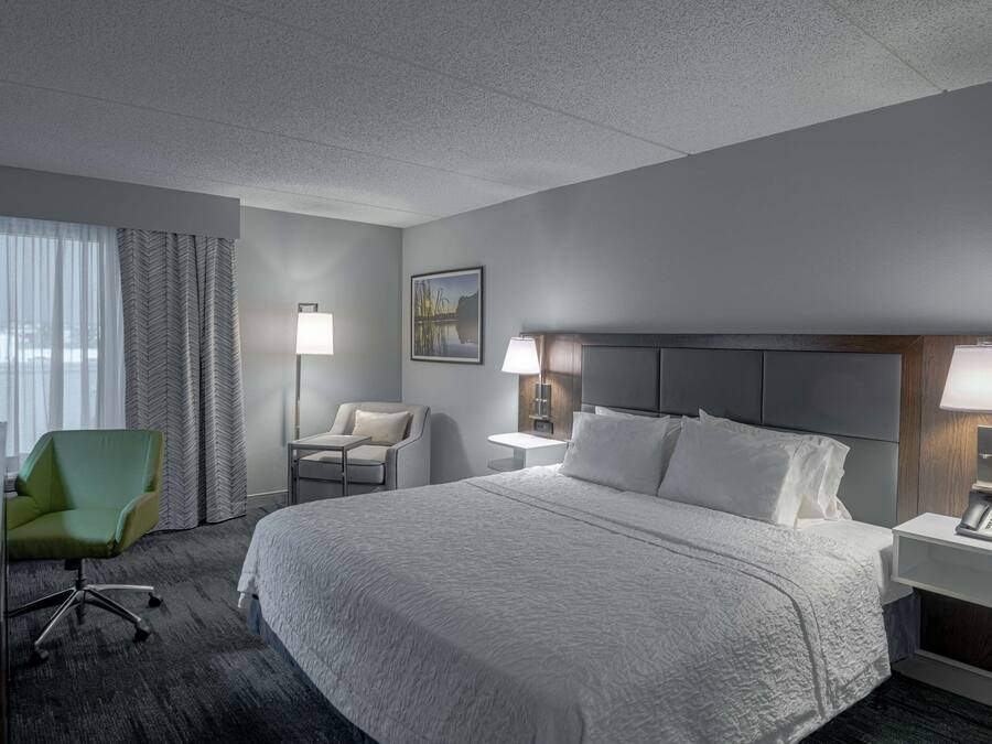A hotel room with a large bed, two bedside tables with lamps, a green chair, a desk, and a window with curtains.