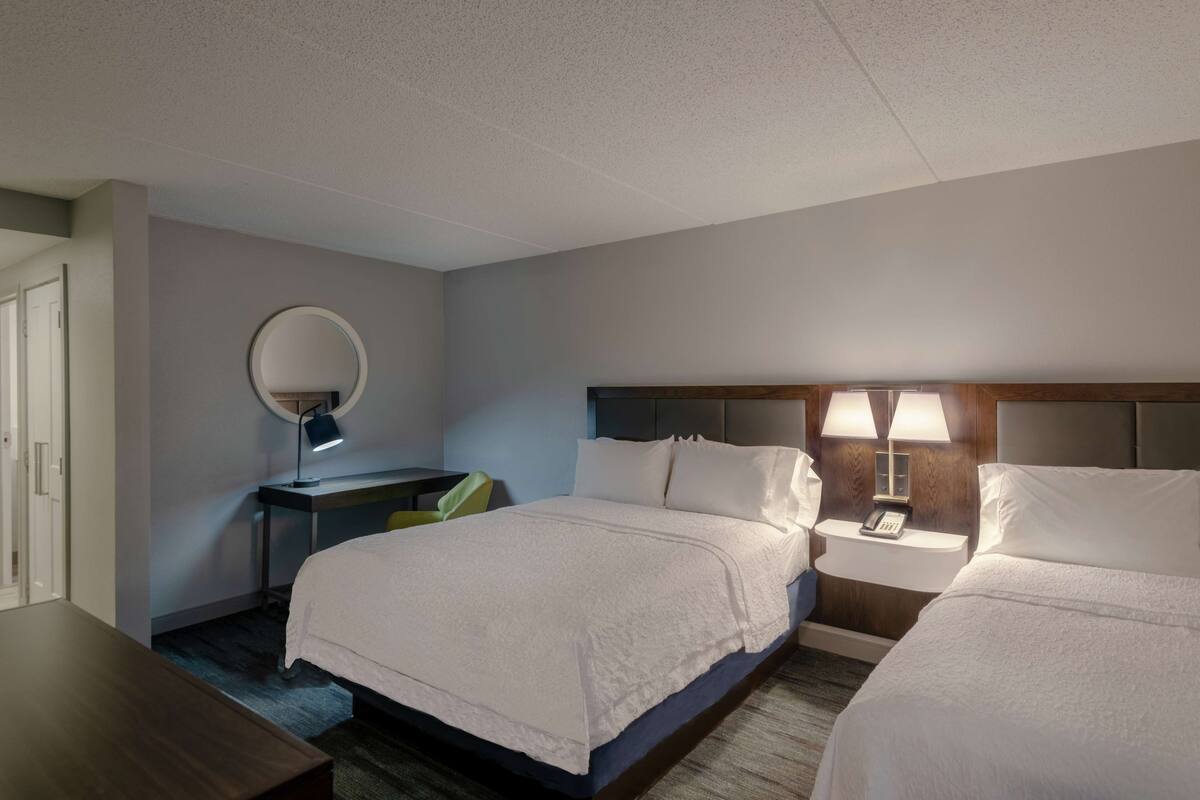 A hotel room with two beds, a desk with a chair, a round mirror, and bedside lamps.
