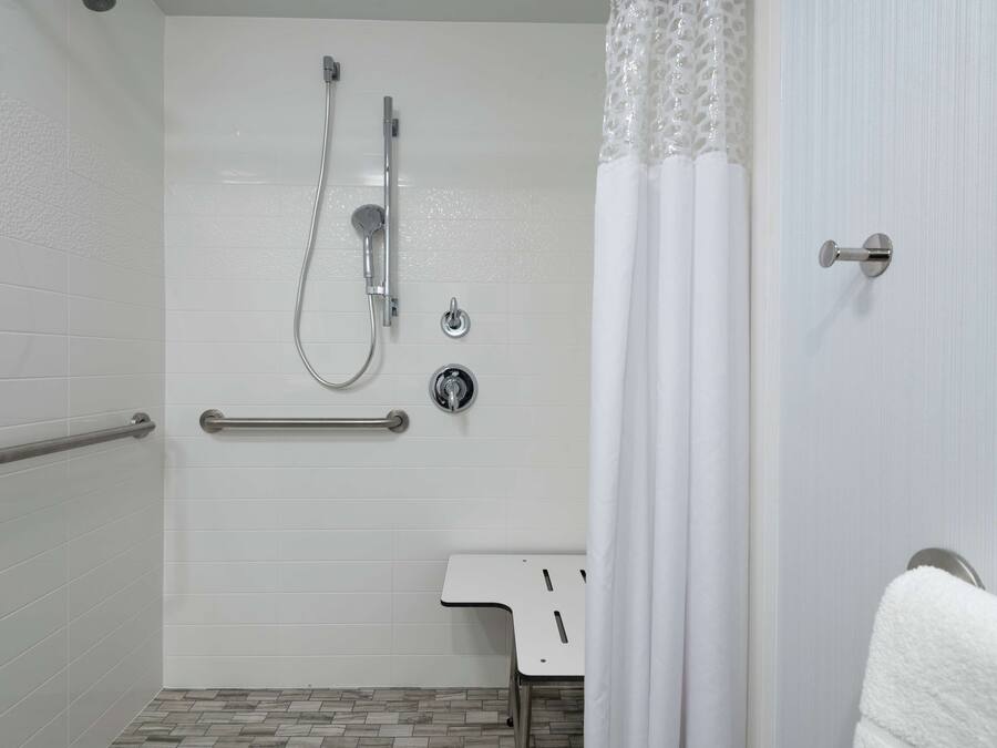 Ein Badezimmer mit weißen Fliesen, einer Dusche inklusive Handbrause, einer Haltestange und einem weißen Waschbecken mit einem Handtuch an der Wand.