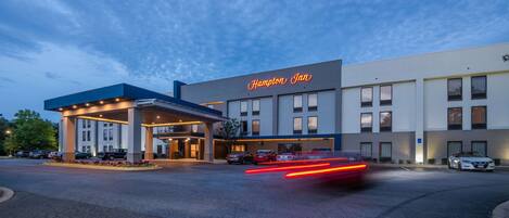 A Hampton Inn hotel with a modern design, featuring a prominent neon sign, a covered entrance, and a parking lot with cars.