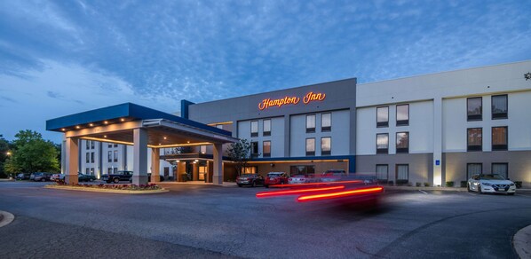 Een Hampton Inn hotel met een modern design, met een opvallend neonbord, een overdekte ingang en een parkeerplaats met auto's.