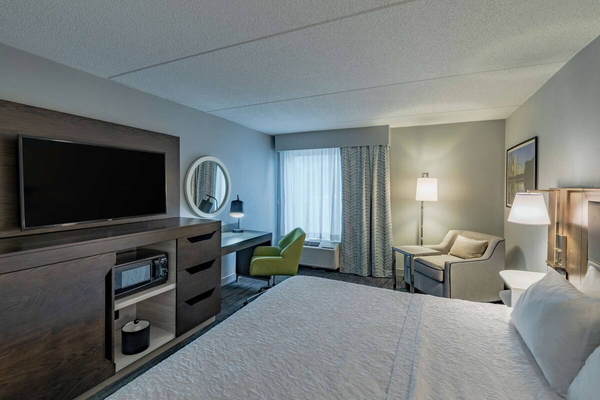 A hotel room with a large bed, a flat-screen TV, a desk with a lamp, a chair, and a window with curtains.