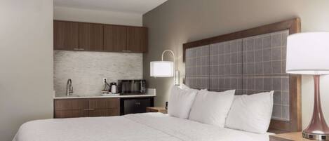 A hotel room with a large bed, a bedside table with a lamp, a small desk with a coffee maker, and a kitchenette with cabinets and a microwave.