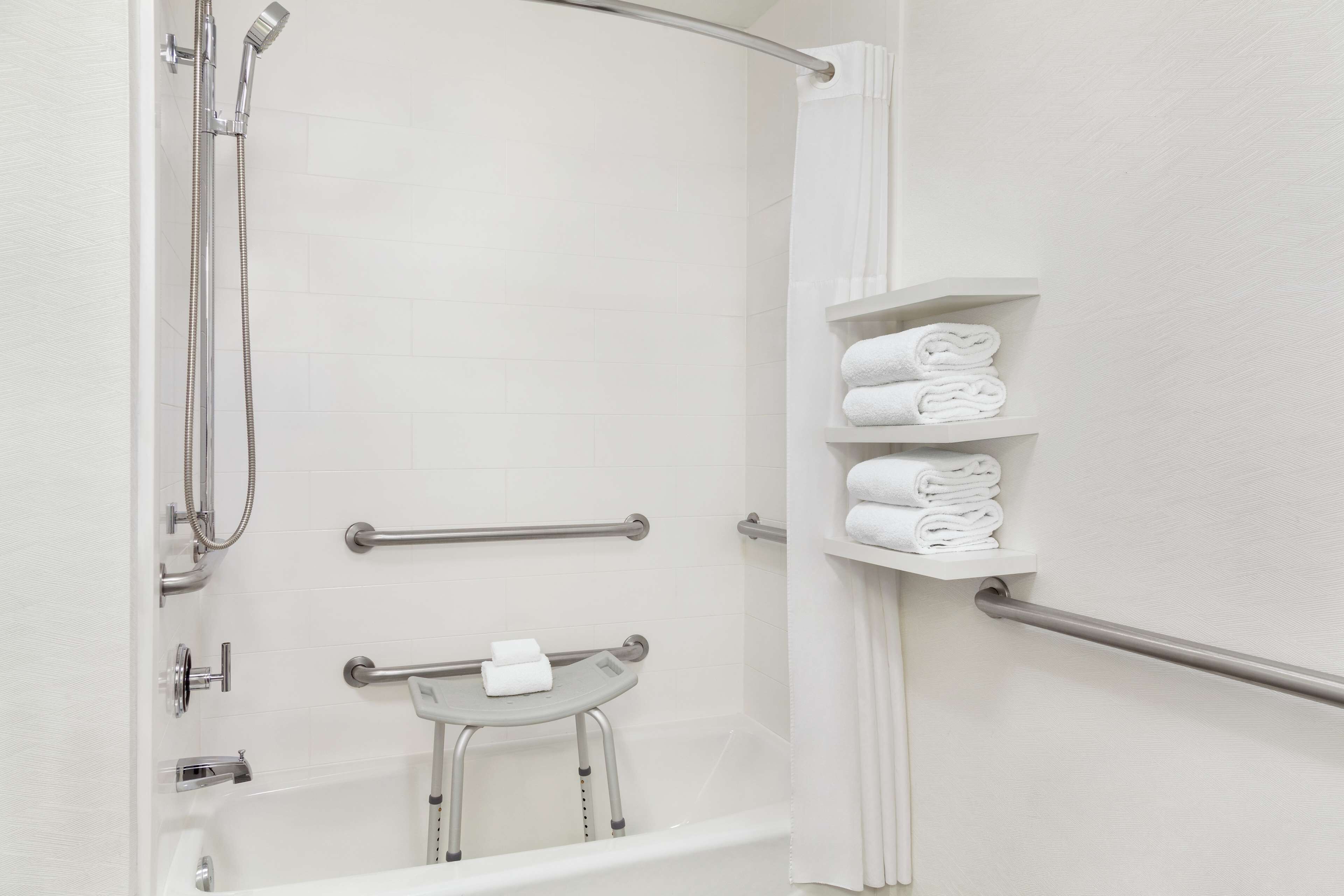 A modern bathroom with a bathtub, a shower, and a handrail. There are shelves with folded towels.