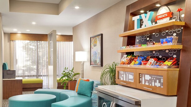 Un hall de hotel con asientos color turquesa, un bar de aperitivos y un ventanal con cortinas translúcidas.