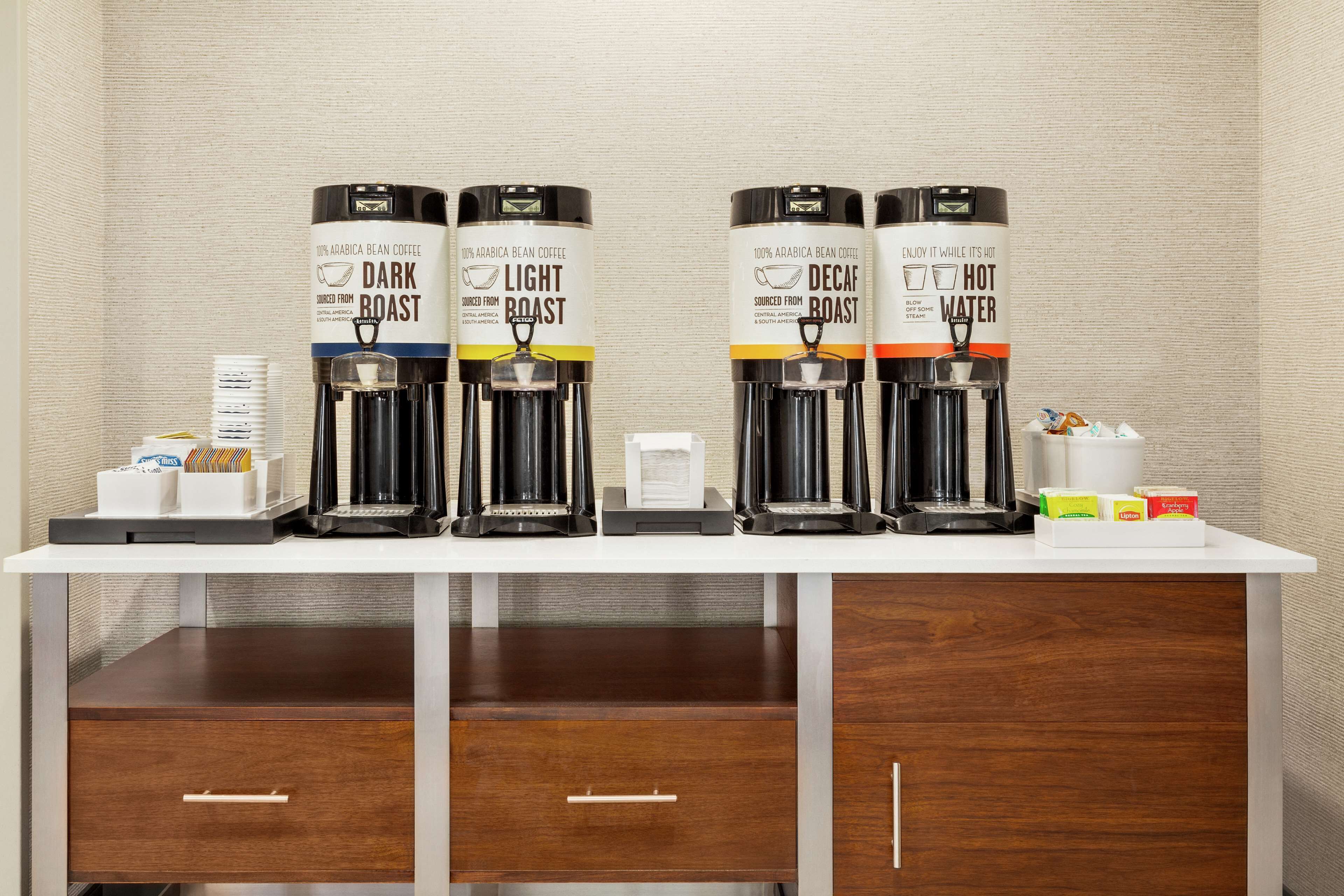 Een koffiehoek met vier koffieautomaatjes, gelabeld 'Dark Roast', 'Light Roast', 'Decaf' en 'Warm Water'. Er staan koffiekapsules en een klein bakje op het aanrecht.
