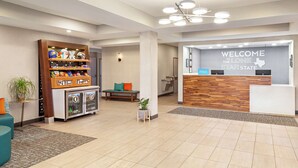 Zona de recepción de hotel con un mostrador de madera, un letrero en la pared que dice “WELCOME THE LONE STAR STATE” y un estante con diversos objetos, incluyendo cascos y un refrigerador pequeño.