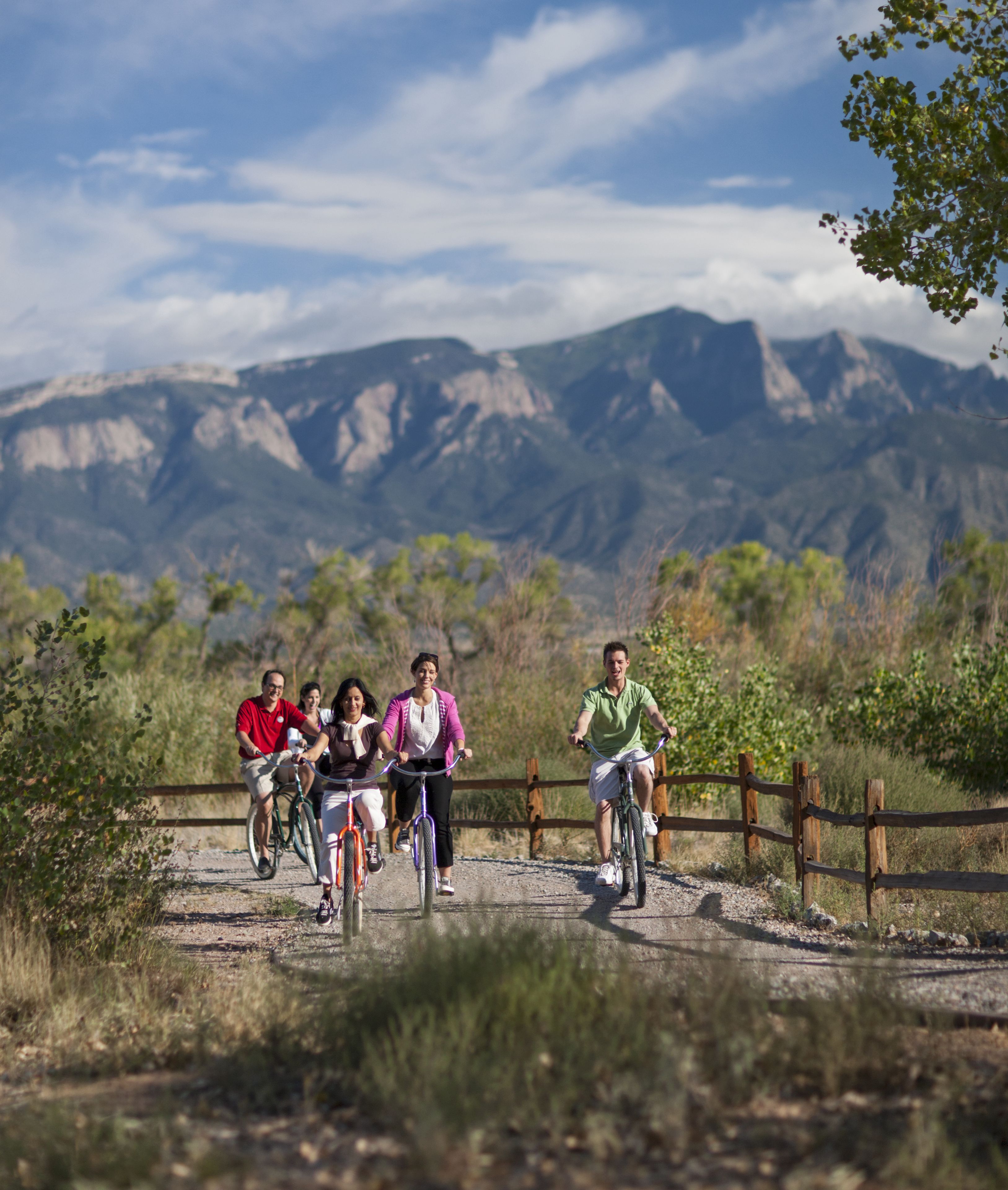 A group of people riding bicycles on a dirt path with a wooden fence and mountains in the background.