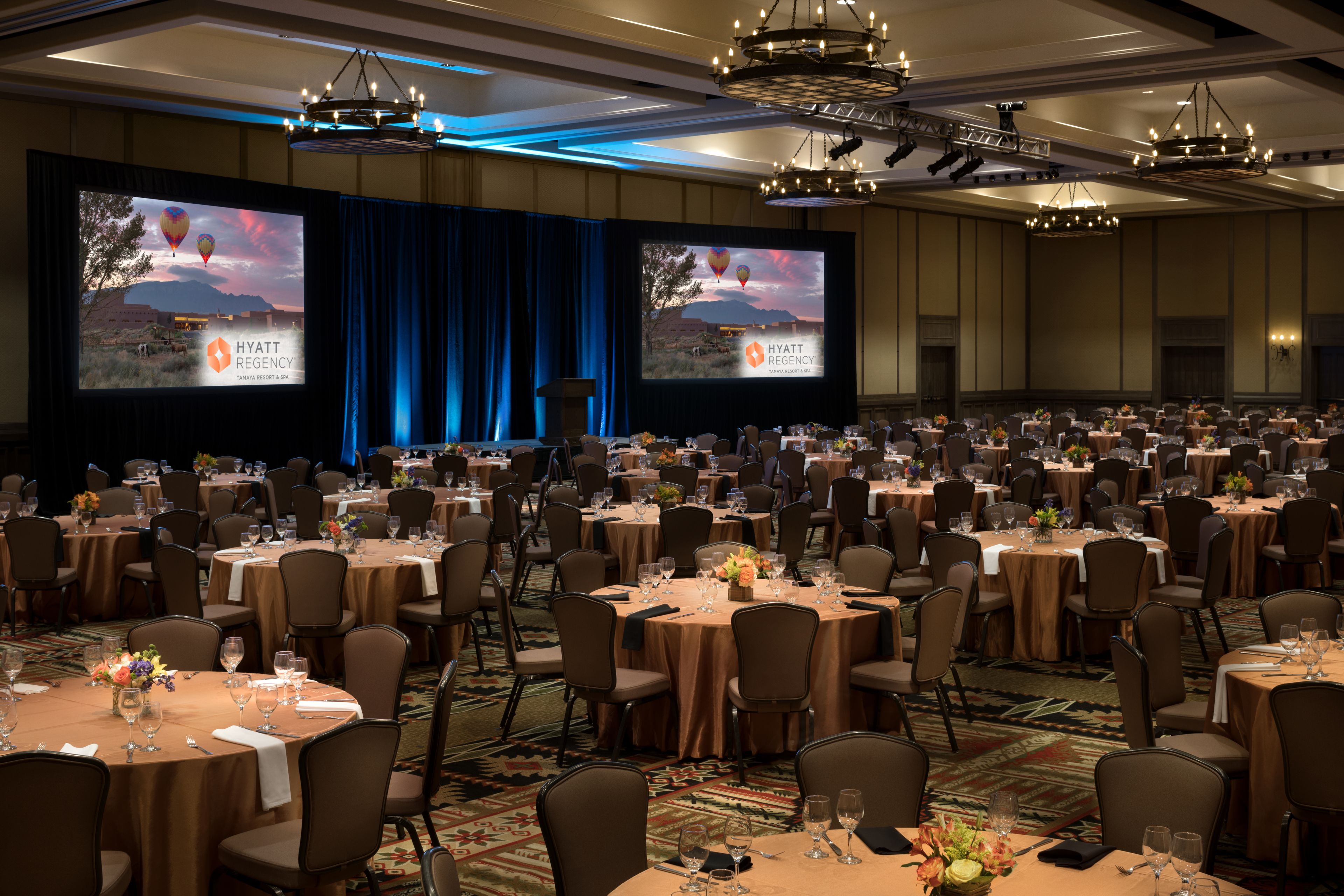 Un amplio salón de banquetes con mesas redondas, sillas y un escenario con una pantalla que muestra el logotipo de Hyatt Regency.