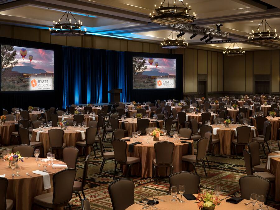 A large banquet hall with round tables, chairs, and a stage with a screen displaying the Hyatt Regency logo.