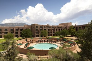 A hotel with a circular pool surrounded by lounge chairs and umbrellas, set against a backdrop of mountains and a clear sky.