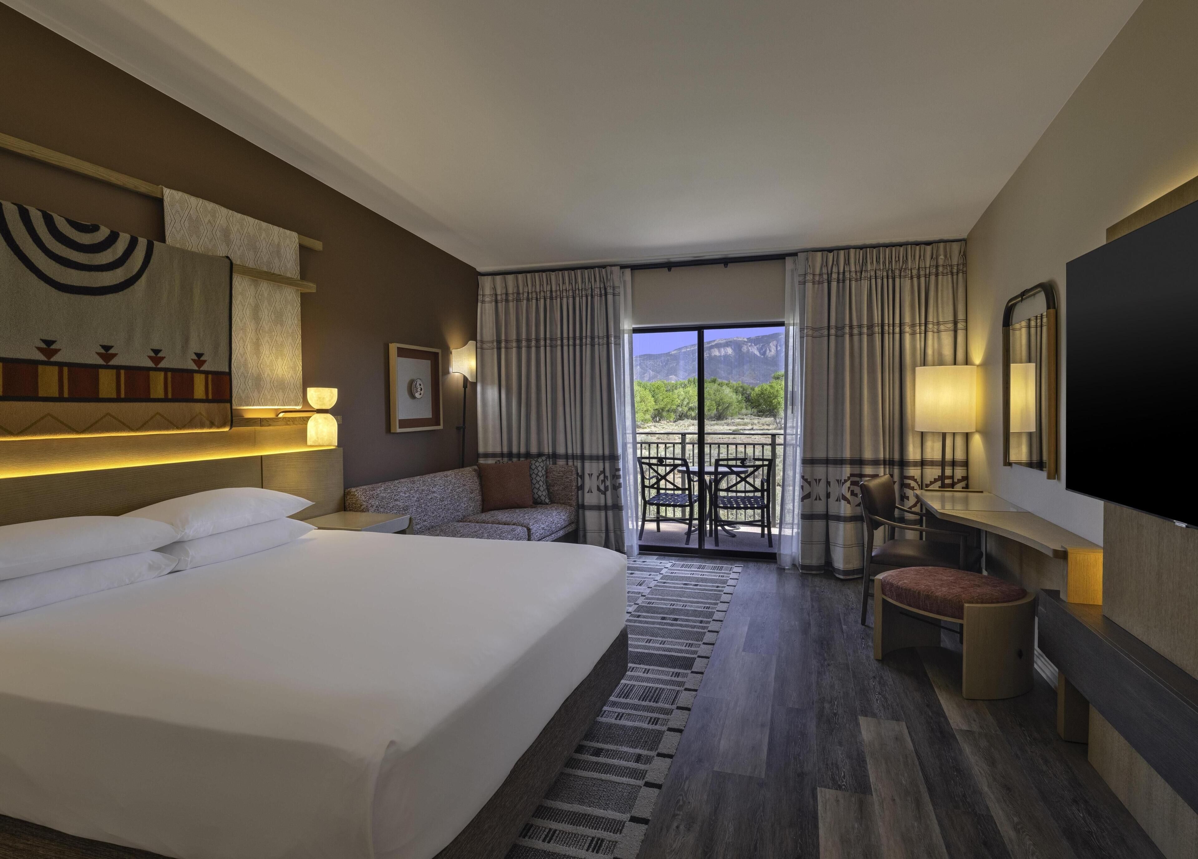 Quarto de hotel com uma cama grande, um sofá, uma mesa pequena, uma televisão e varanda com vista para as montanhas.