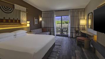 Quarto de hotel com uma cama grande, um sofá, uma mesa pequena, uma televisão e varanda com vista para as montanhas.