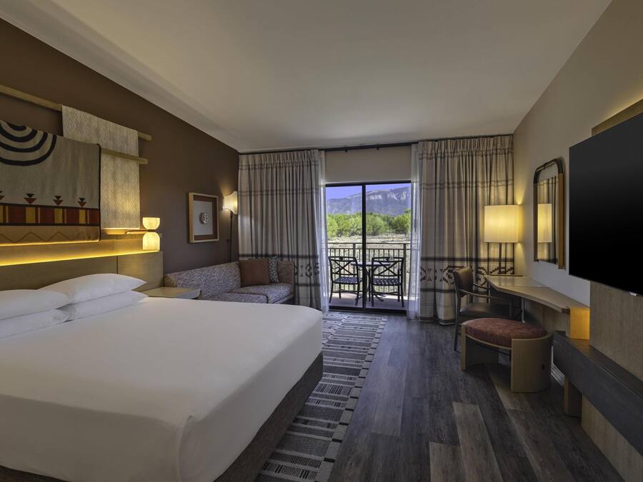 Quarto de hotel com uma cama grande, um sofá, uma mesa pequena, uma televisão e varanda com vista para as montanhas.