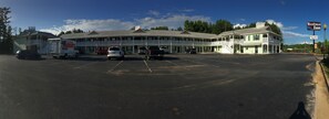 Un motel con un edificio central y una estructura más pequeña adosada, un estacionamiento con varios vehículos, y un cielo despejado.