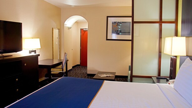 A hotel room with a bed, a desk with a lamp, a television, a chair, and a door leading to another room. - Super 8 by Wyndham Irving/DFW Apt/North (Irving)