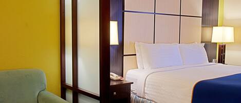 Habitación de hotel con cama, mesita de noche con lámpara, una silla y un teléfono sobre la mesa.