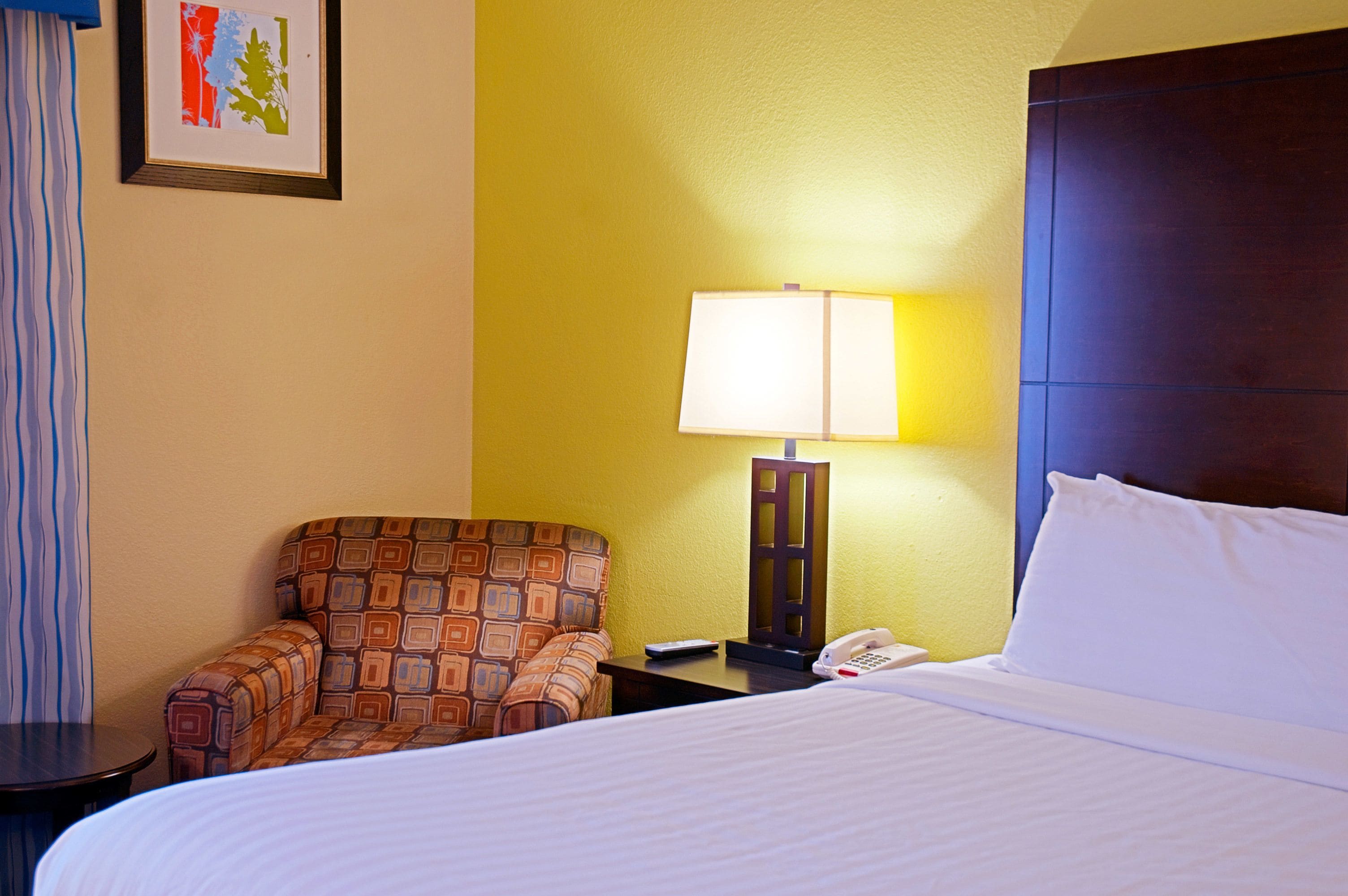 A hotel room with a bed, a patterned armchair, a bedside table with a lamp, and a telephone.
