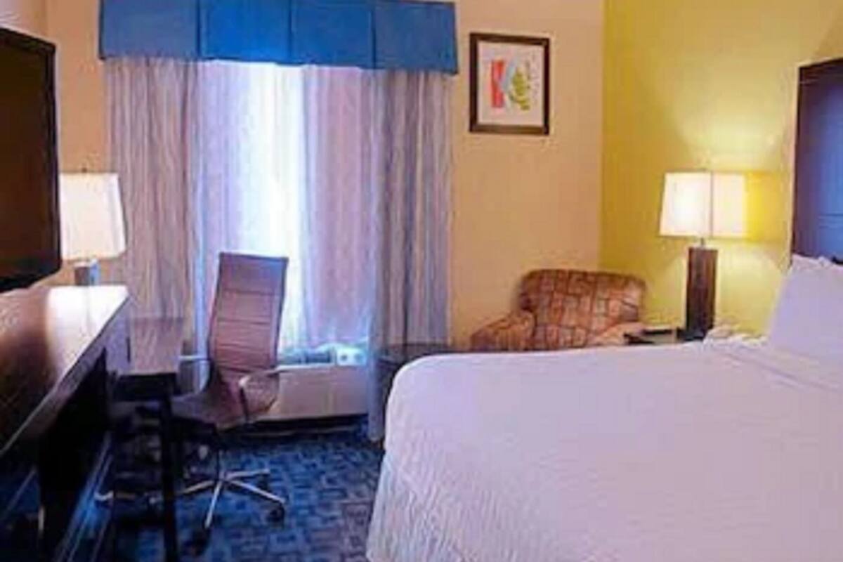 A hotel room with a bed, a desk, a chair, a lamp, and a window with curtains.