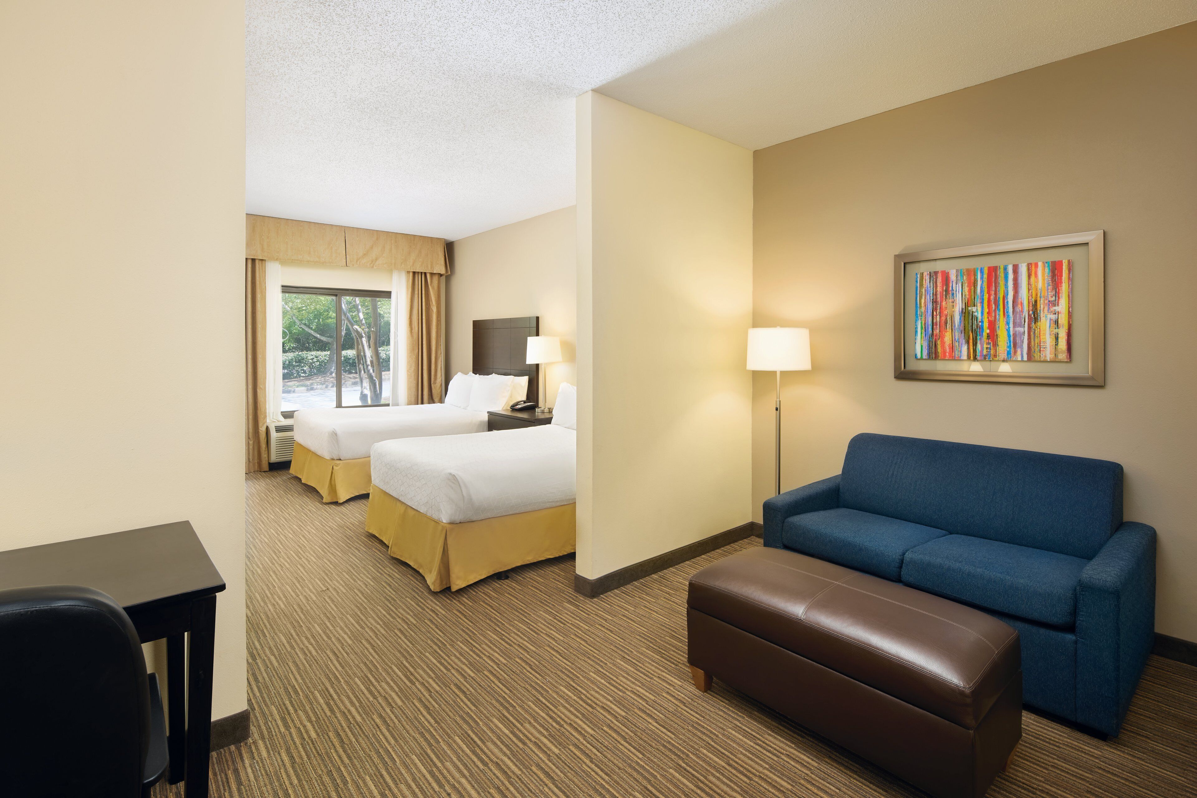 A hotel room with two beds, a blue sofa, a brown ottoman, a desk, and a framed abstract painting on the wall.