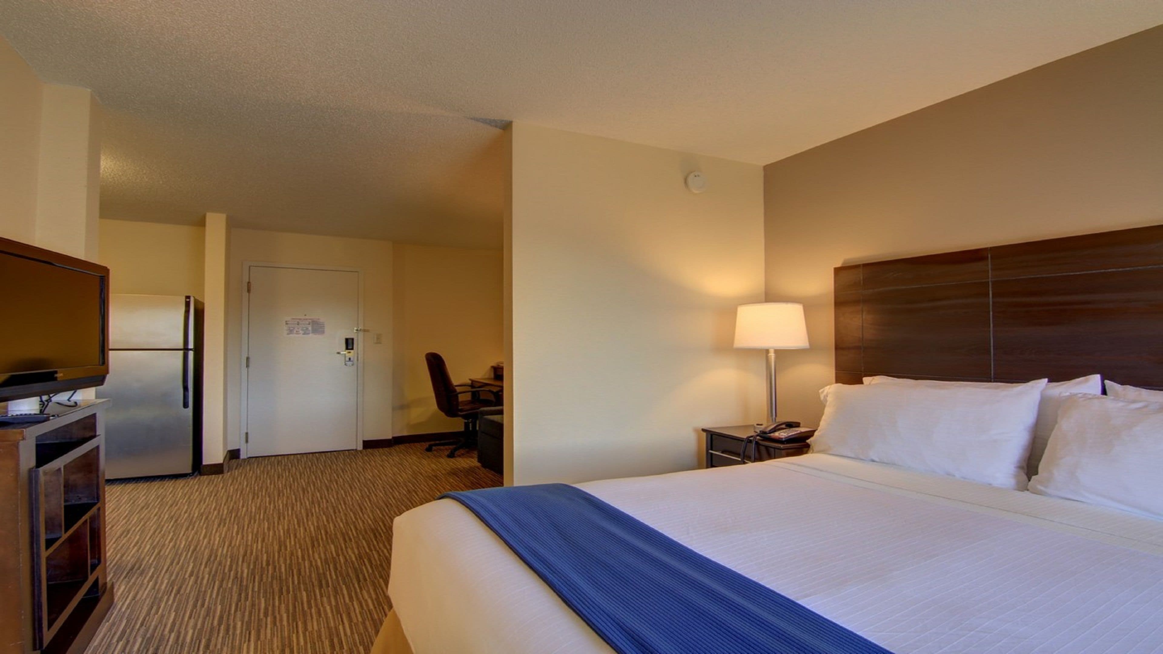 A hotel room with a bed, a desk, a chair, a refrigerator, and a lamp.