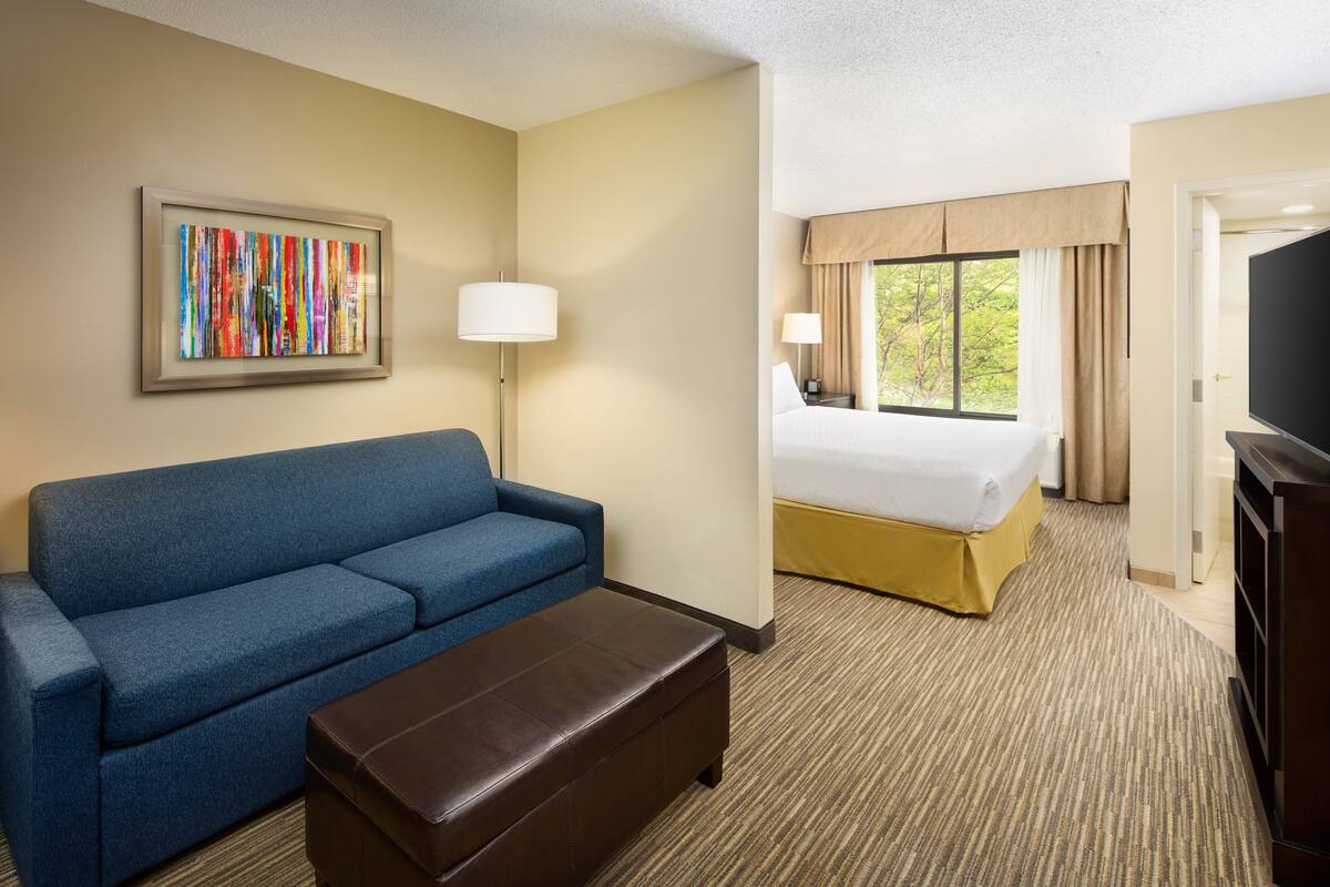 Ein Hotelzimmer mit blauem Sofa, braunem Hocker, Bett mit beigeartigem Kopfteil, großem Fenster mit Vorhängen und einem Flachbildfernseher.