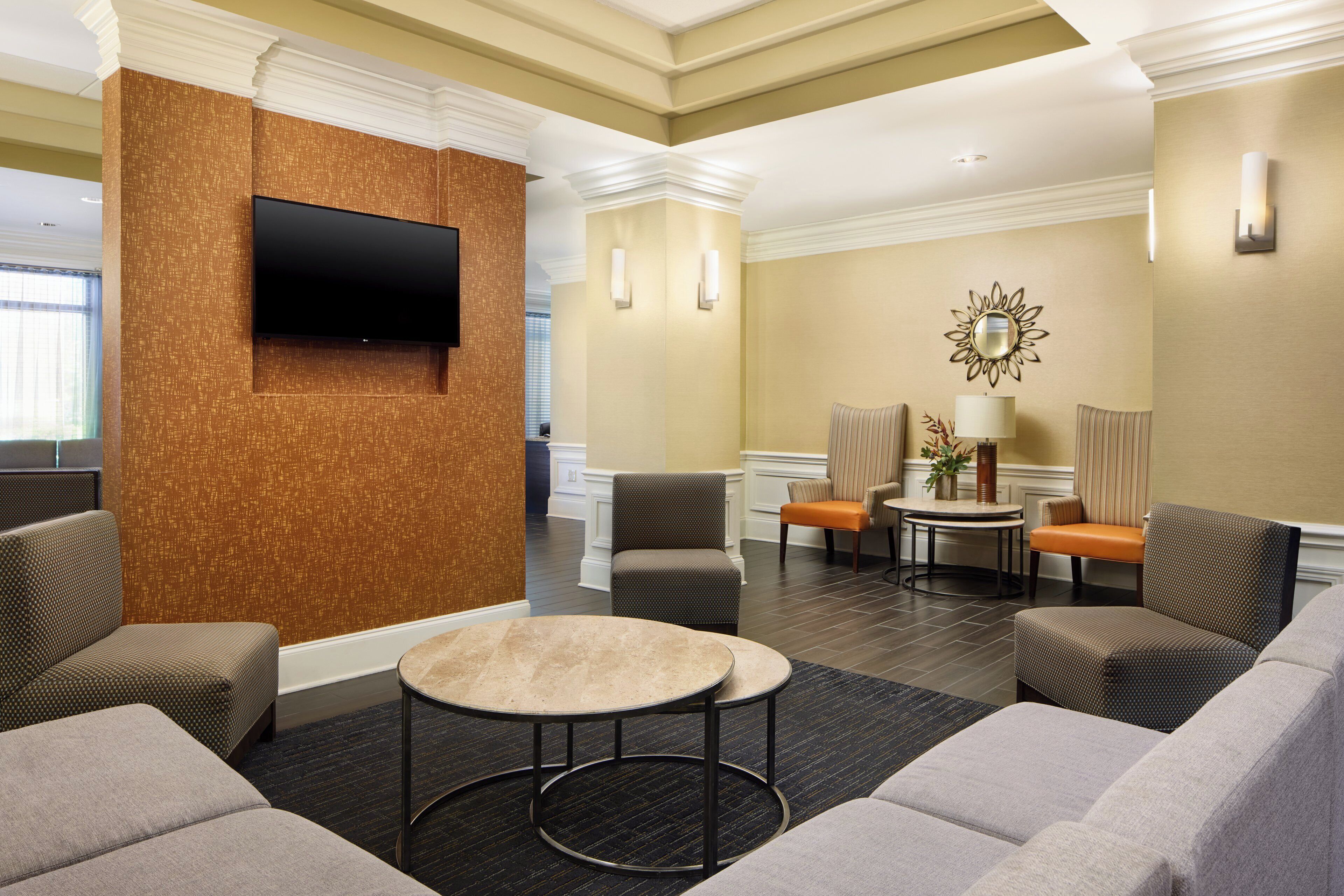 Un vestíbulo de hotel con un televisor de pantalla plana montado en una pared con textura, una mesa de centro redonda y varias opciones de asiento, incluyendo sofás y sillones.