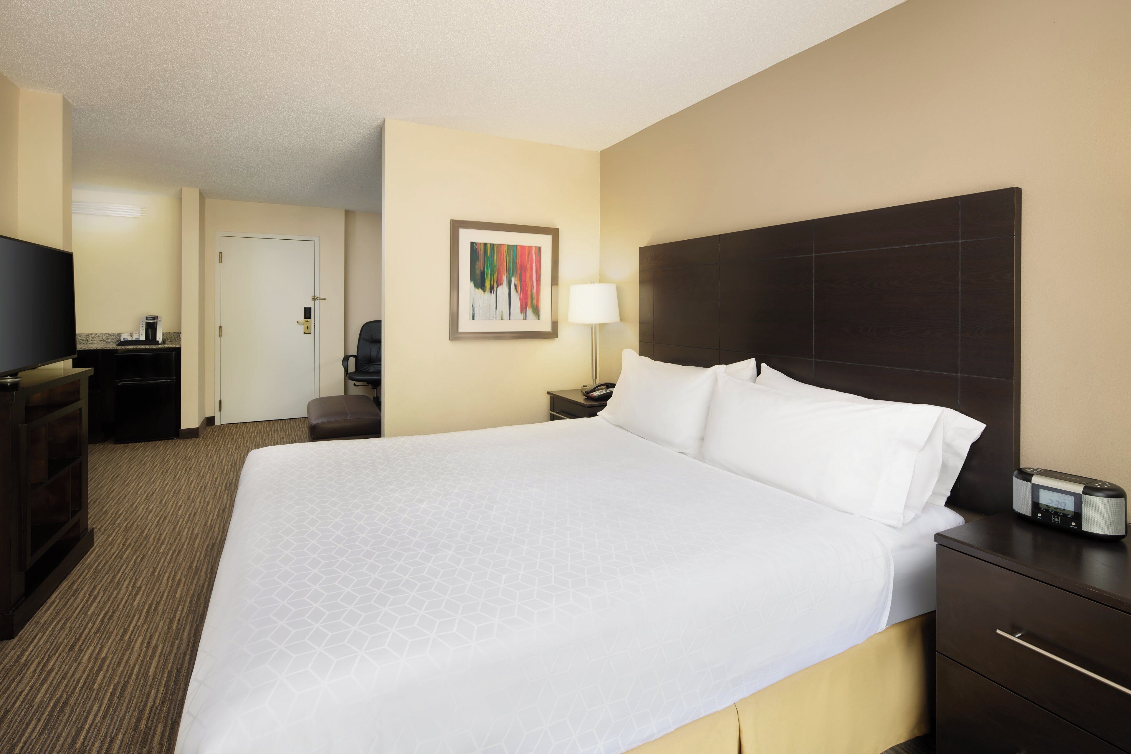 A hotel room with a large bed, a desk with a chair, a TV, a lamp, and a painting on the wall.