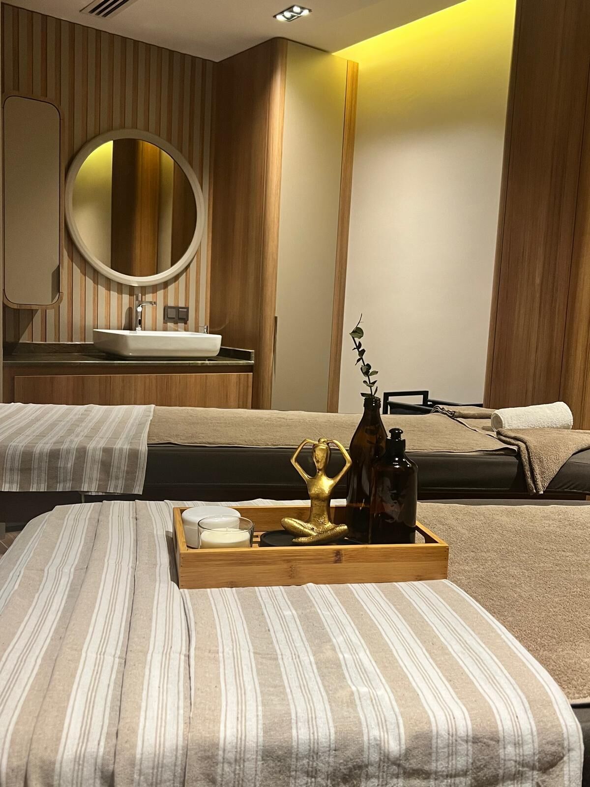 A hotel room with a bed, a bedside table with a tray of toiletries, a round mirror, and a wooden wall paneling.